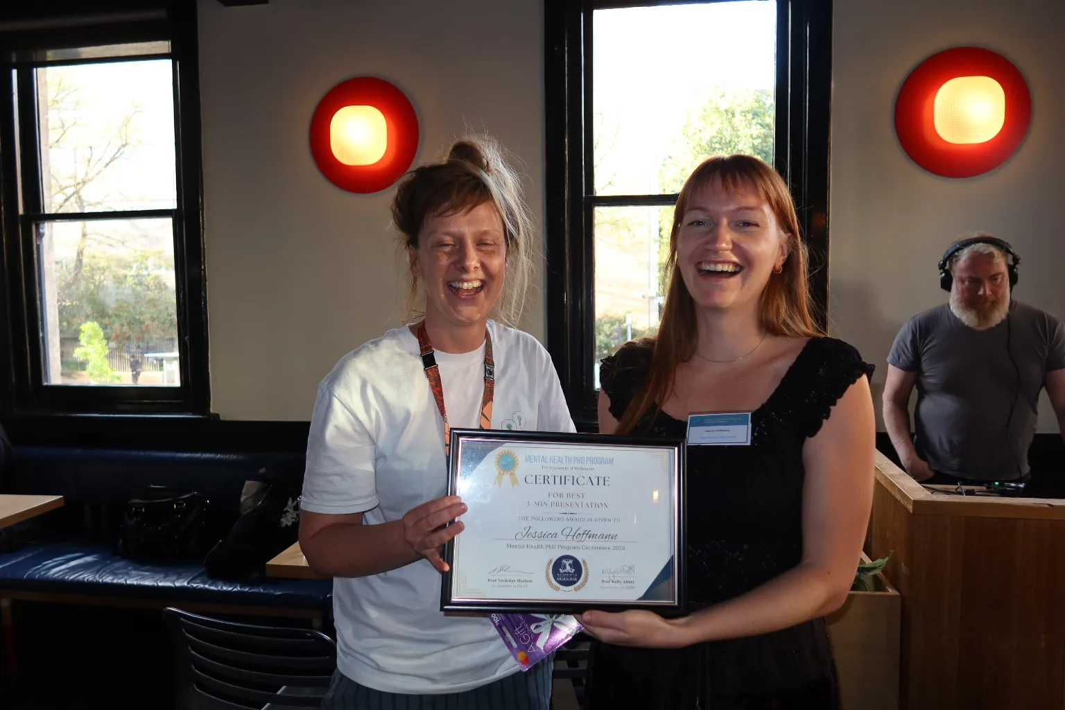 Jessica Hoffmann and another person laugh while holding Jessica’s framed certificate for Best Three-Minute Presentation at the 2026 Mental Health PhD Program Conference